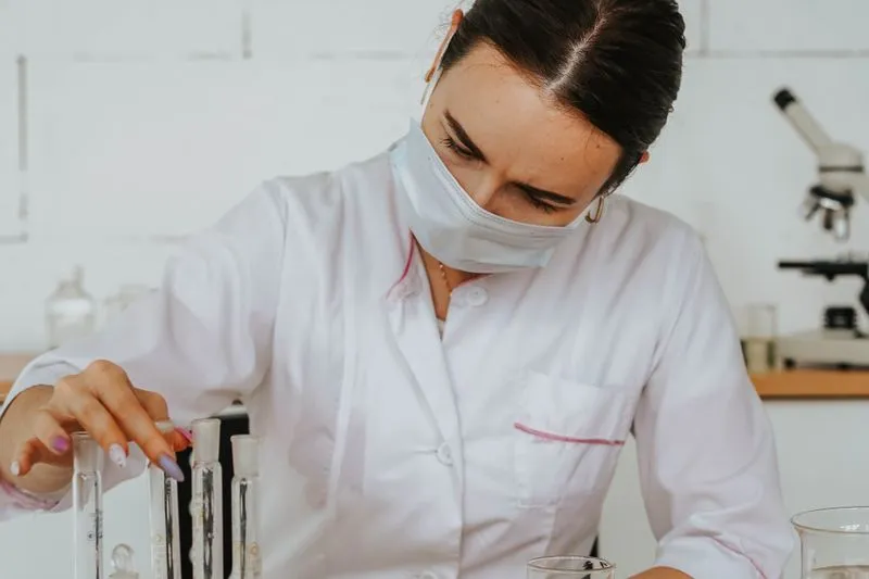 Researcher in a white lab coat and surgical mask handling test tubes, with a microscope in the background