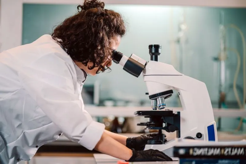 Curly-haired researcher in a white lab coat and black gloves, leaning over the eyepiece of a microscope