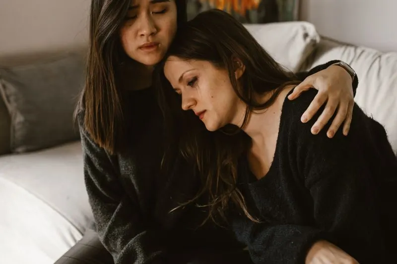 Woman in tears resting her head on a friend's shoulder as the friend holds her close on a sofa
