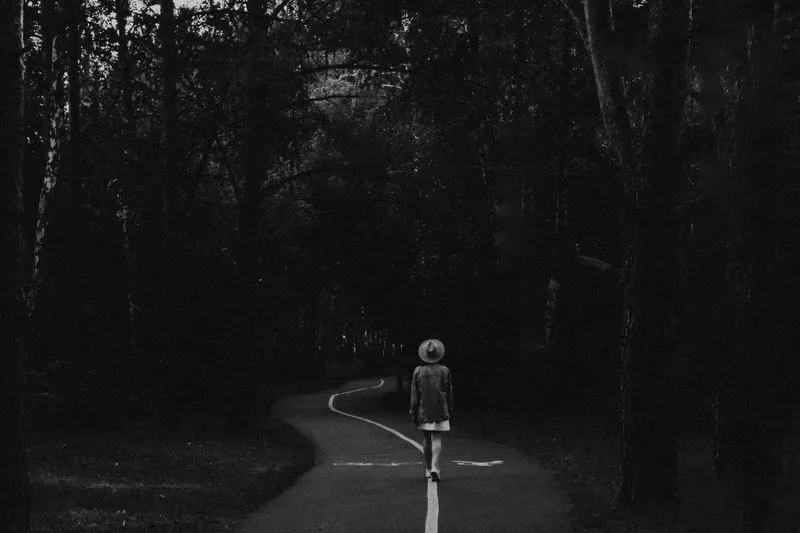 Woman in a hat walking alone down a winding path through a dark forest, seen from behind, in black and white