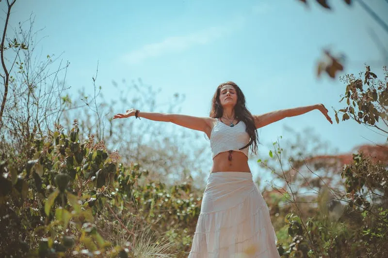 Woman with her arms spread wide and eyes closed, standing among greenery under a blue sky