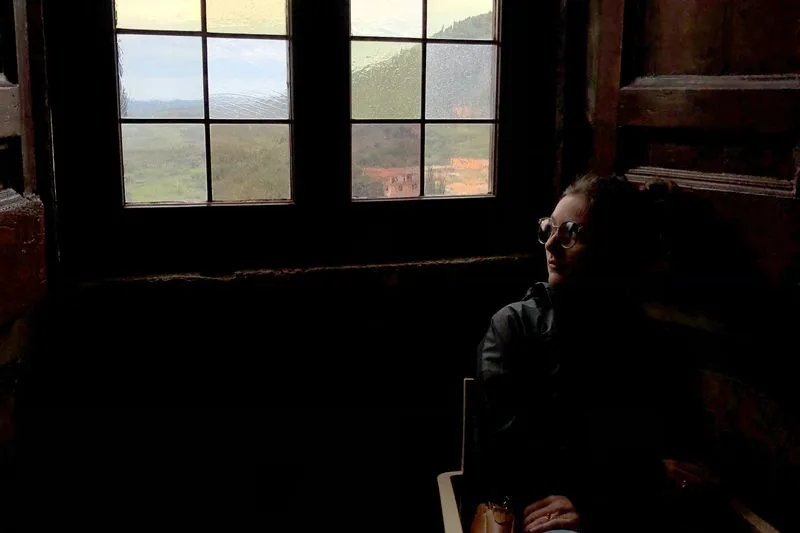 Young woman in glasses sitting in a dim room, gazing towards a window that looks out over green hills