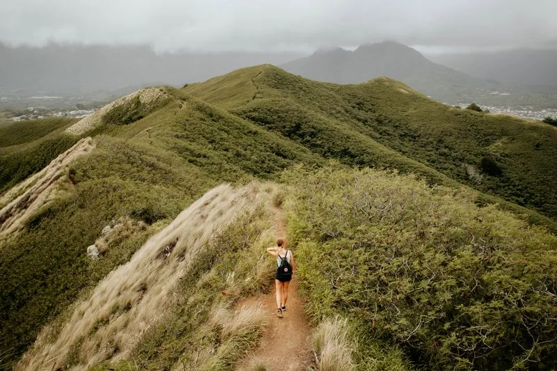 Hiker walking along a ridge trail that winds across green hills under a cloudy sky