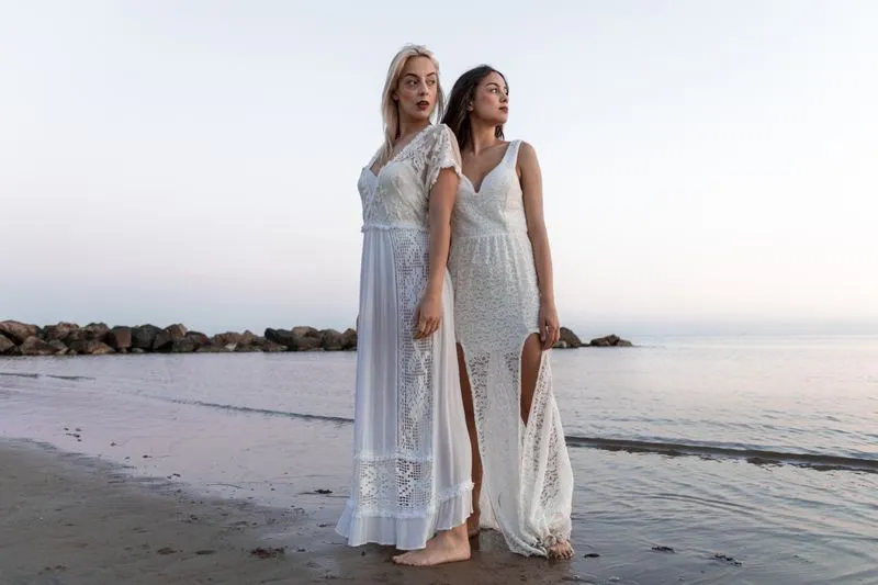 Two barefoot women by the sea in long white dresses with crochet and lace panels