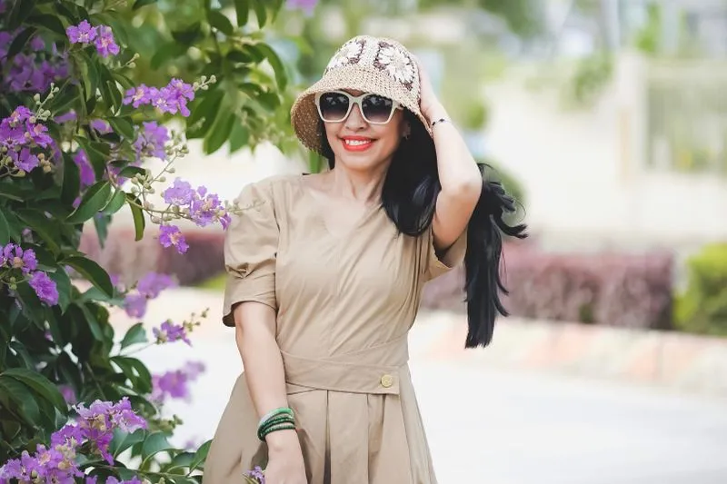 Smiling woman in a beige dress, crochet bucket hat and sunglasses, beside purple blossoms