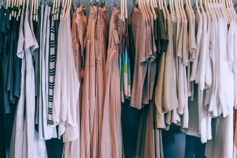 Flowing summer clothes in light tones, including pleated terracotta-pink dresses, on wooden hangers