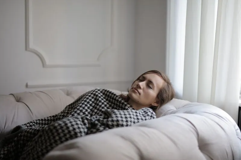 Woman lying on a sofa under a houndstooth throw, eyes closed, exhausted