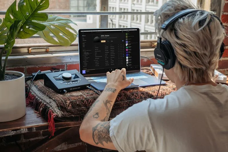 Person seen from behind, wearing over-ear headphones, browsing a track list on their laptop
