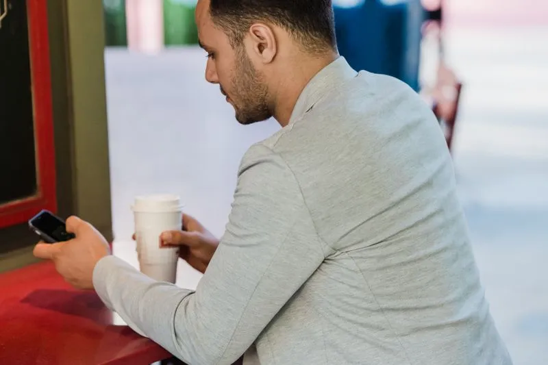 Man at an outdoor café table, phone in one hand and a coffee cup in the other