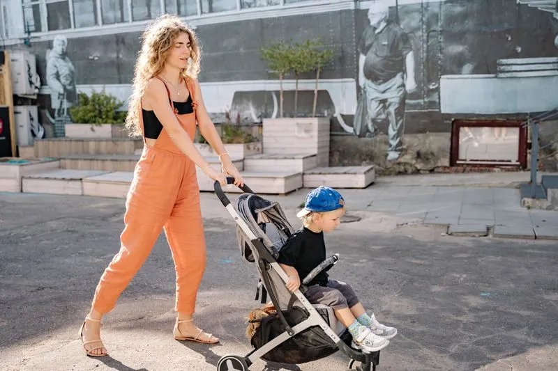Young woman in an orange jumpsuit pushing a buggy with a little boy sitting in it, on a sunny street