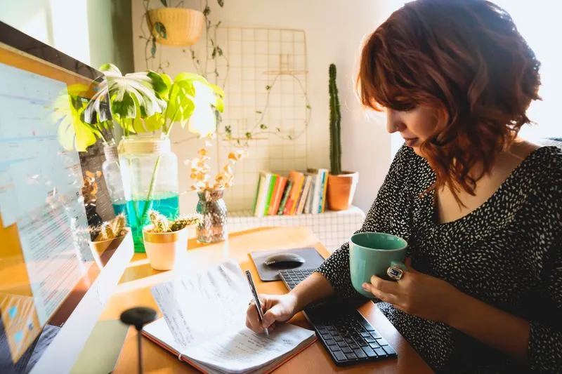 Woman with auburn hair writing in a notebook at her desk, holding a green mug