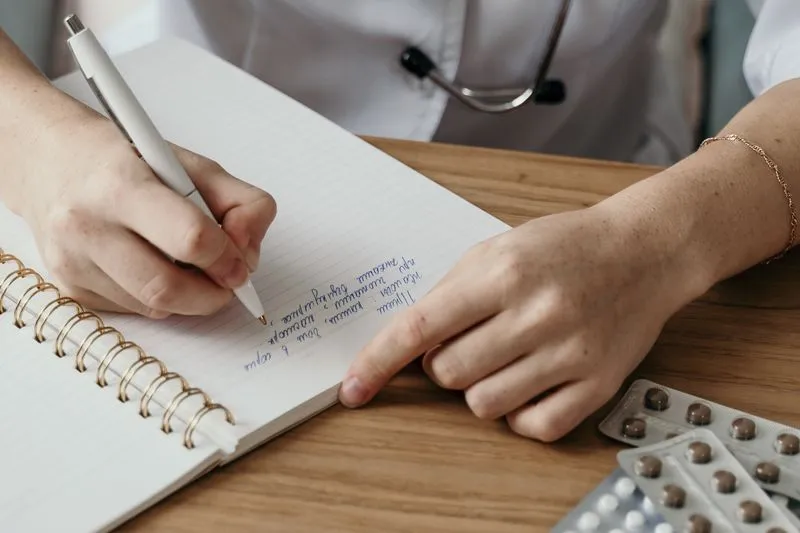 Doctor in a white coat taking notes at her desk, next to blister packs of tablets