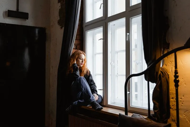 Woman sitting alone on a windowsill, knees drawn up, looking away