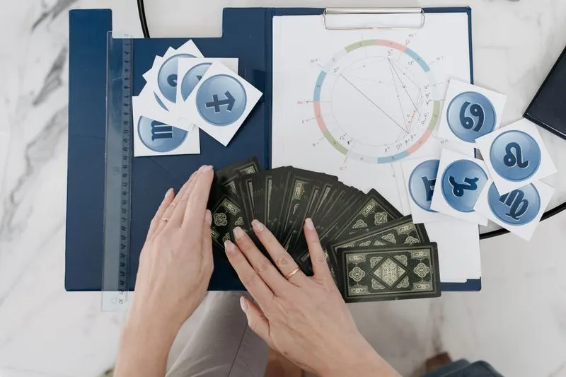 Hands fanning out tarot cards in front of a printed birth chart on a clipboard, surrounded by zodiac sign cards