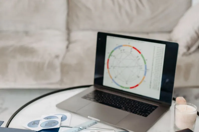 Laptop on a coffee table showing a birth chart wheel, next to zodiac sign cards, a ruler and a candle