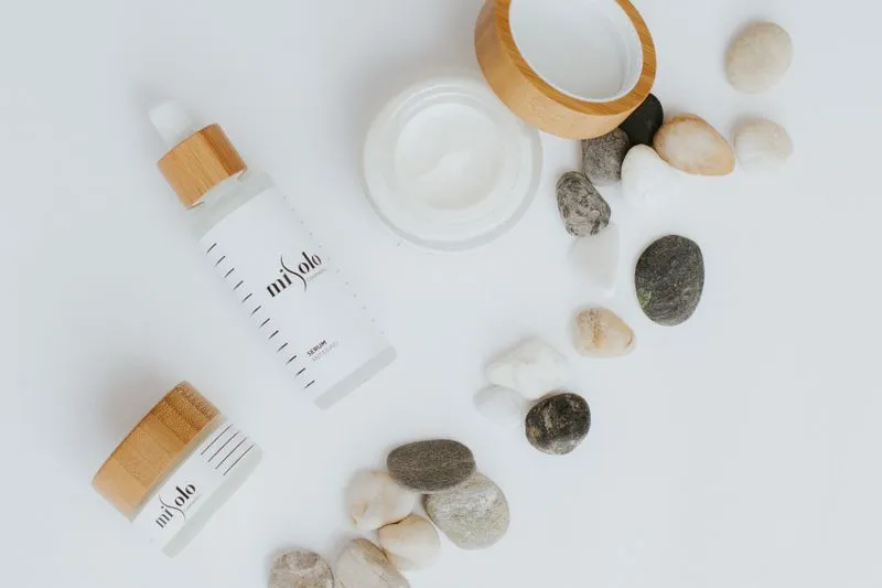 Dropper bottle of serum and an open jar of white cream with a bamboo lid, among pebbles on a white background