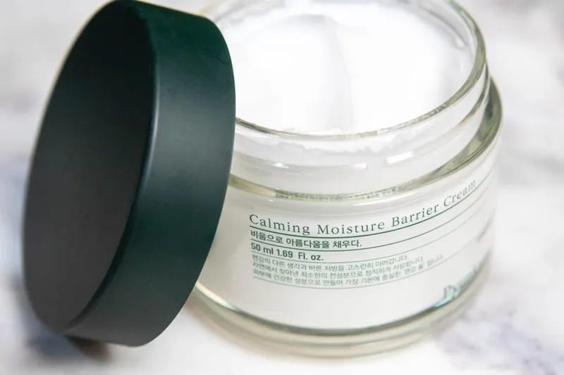 Open jar of calming barrier cream, thick white texture, on marble