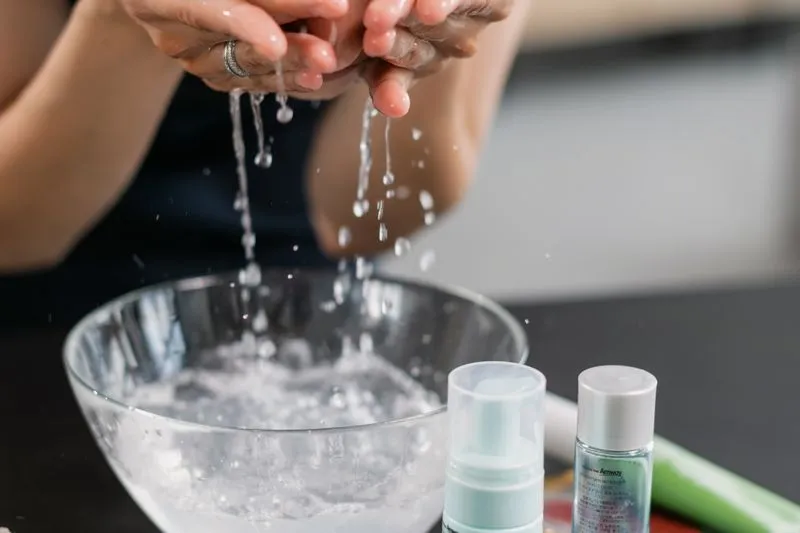 Hands splashing water over a glass bowl, with two small skincare bottles beside it