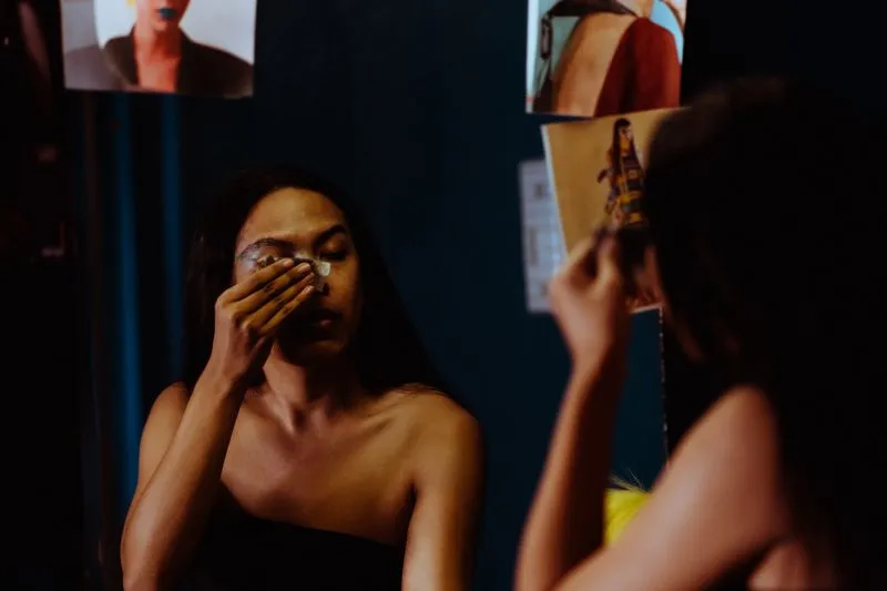 Young woman removing eye make-up with a cotton pad in front of a mirror in a dim room