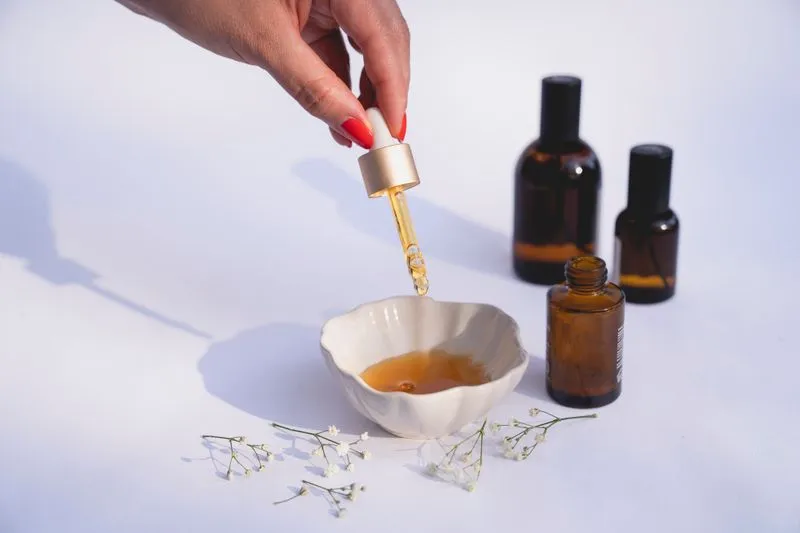 Hand holding a dropper of golden serum over a small dish, amber bottles in the background