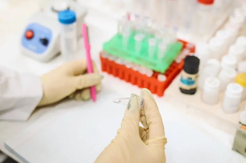 Gloved hands holding a small tube in a laboratory, racks of test tubes in the background