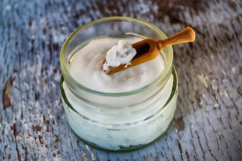 Glass jar filled with solid coconut oil, a small wooden scoop resting on top, on a weathered wooden board