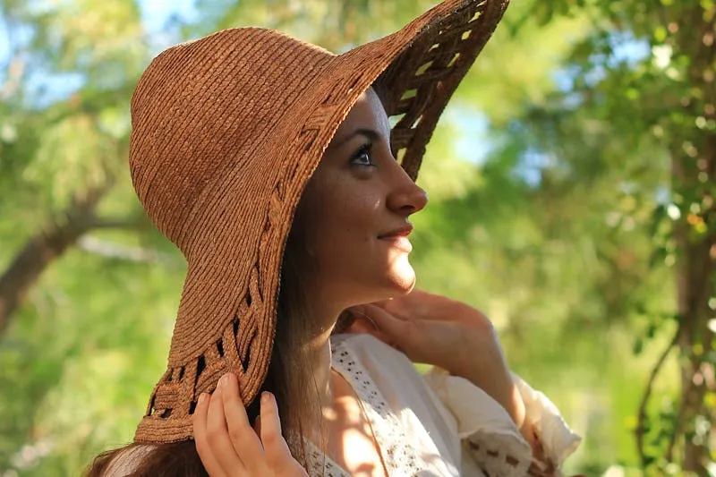 Young woman in a wide-brimmed straw hat, her face in the sun beneath trees