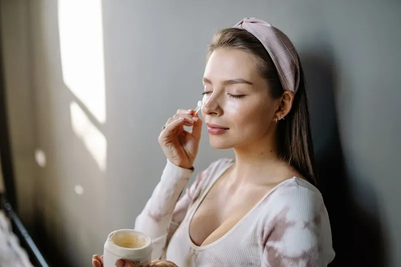 Young woman with her eyes closed, wearing a pink check headband, applying cream from a jar with a small spatula, in window light
