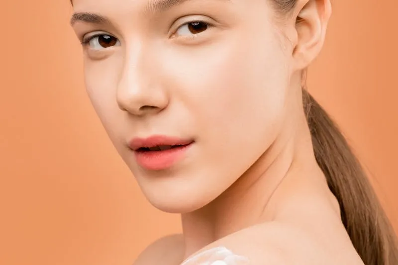 Young woman with her hair tied back, looking over her shoulder, a dab of cream on her shoulder, orange background