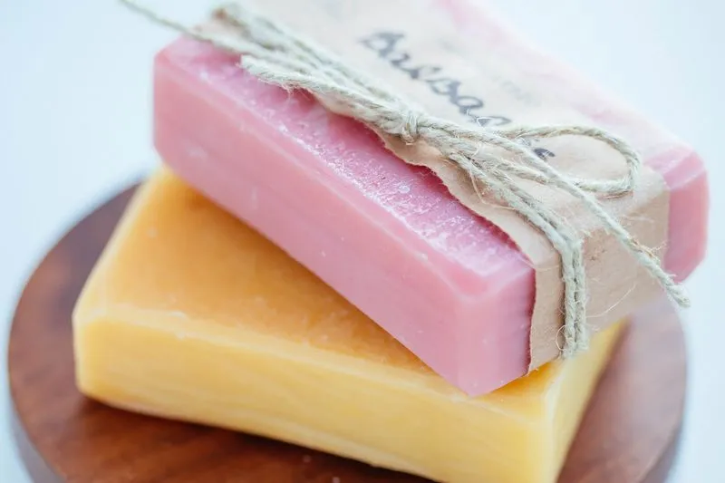 Two handcrafted soap bars, pink and yellow, stacked and tied with twine, on a wooden dish