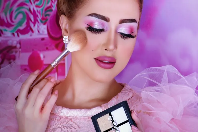 Woman in pink evening make-up sweeping a large powder brush over the top of her cheekbone, holding a palette of pale powders