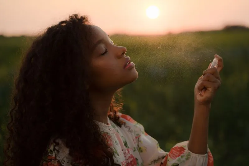 Young woman with curly hair spraying a mist near her face, eyes closed, in a field at sunset