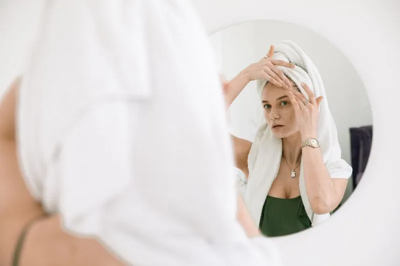 Woman with her hair wrapped in a towel, working moisturiser into her forehead with her fingertips in front of a round mirror