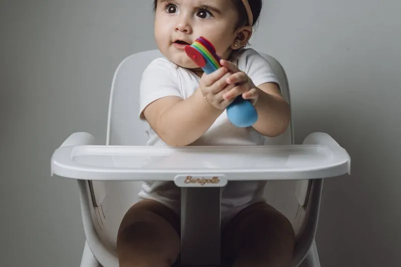 Baby girl sitting upright in a high chair, holding a toy in both hands near her mouth