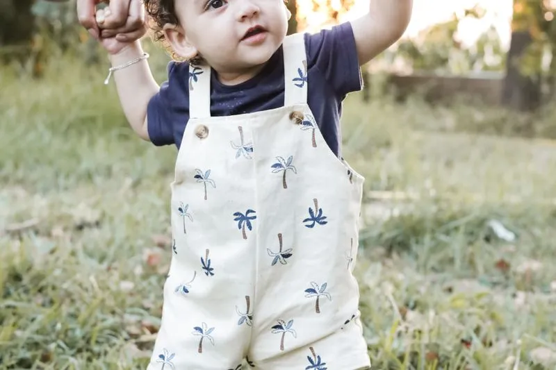 Baby in dungarees walking on the grass, arms raised, held by an adult