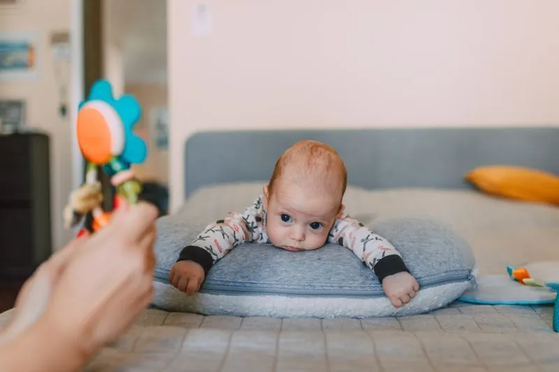 Baby lying on their tummy on a cushion, chin resting on the cushion, watching a rattle held out by an adult