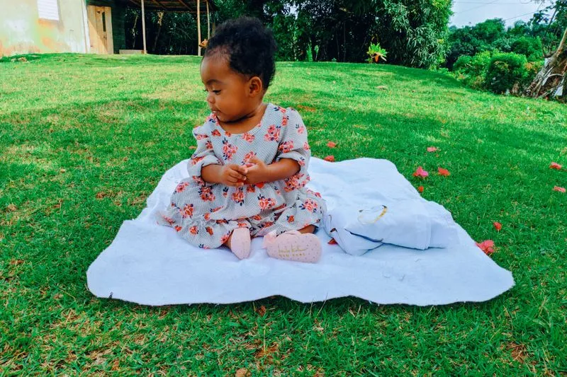 Baby girl sitting unsupported on a blanket spread on the grass, hands clasped