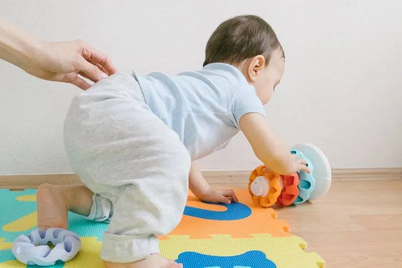 Baby on all fours on a foam play mat, reaching for a toy, an adult's hand resting on their back