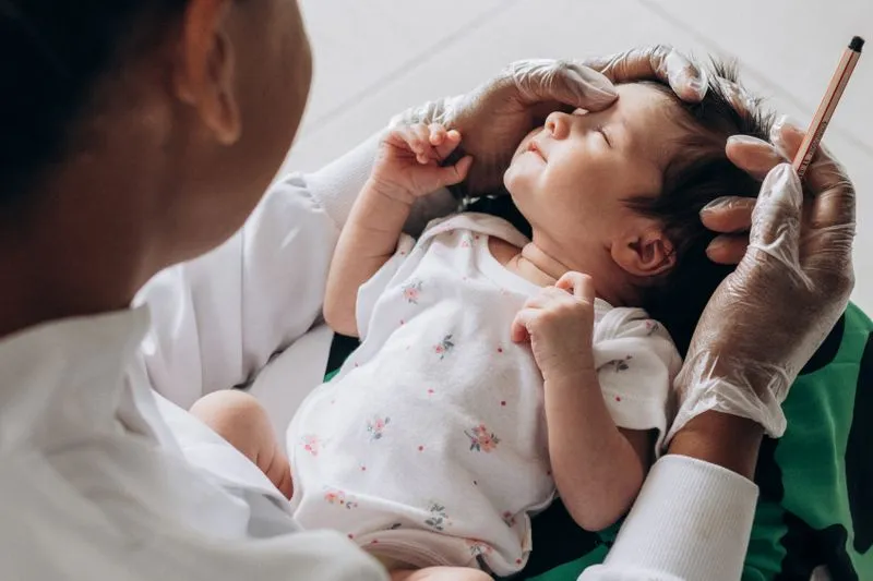 A health professional in a white coat and gloves examining the head of a baby with closed eyes, in a white bodysuit with small flowers