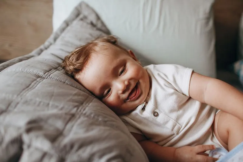 Baby in a white bodysuit laughing, head resting on a grey cushion