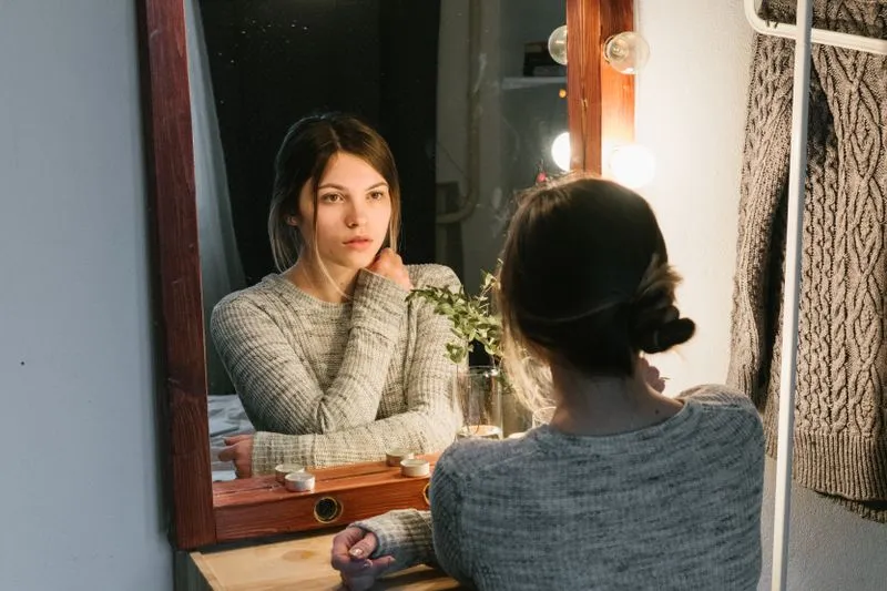 Young woman looking thoughtfully at her reflection in a mirror, chin in hand