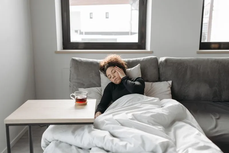 Woman lying on a sofa under a duvet, hand on her temple, a cup of tea beside her