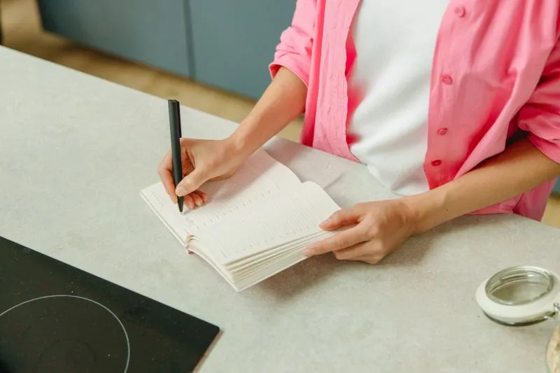 Woman in a pink shirt writing in an open notebook on a kitchen counter