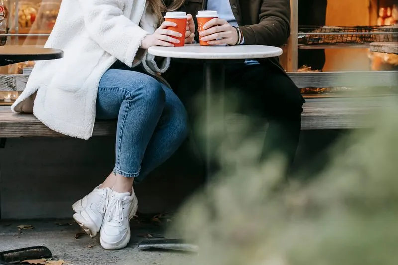 A couple sitting side by side on a bench outside a café, each holding an orange takeaway cup on a small round table