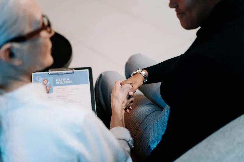 Handshake during an interview between a grey-haired woman holding a CV on a clipboard and a woman in a black jumper