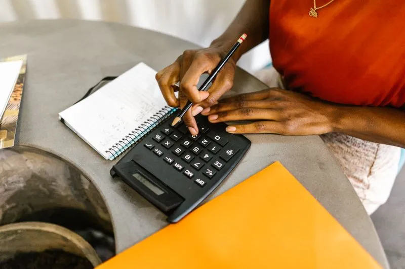 A woman's hands working a calculator with a pencil, next to a notebook and an orange folder