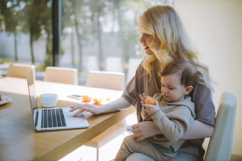 Mother working on a laptop at a dining table, her baby on her lap holding a piece of fruit