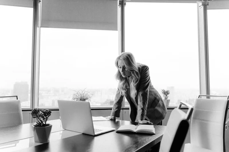 Woman in a blazer leaning over a boardroom table in front of floor-to-ceiling windows overlooking a city, in black and white