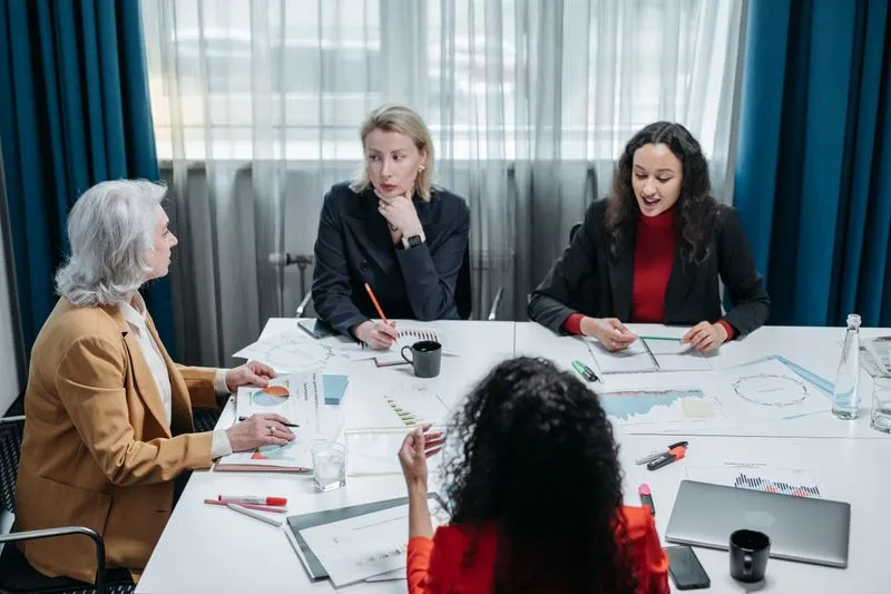 Four women in a meeting around a white table covered with printed charts, notebooks and a laptop