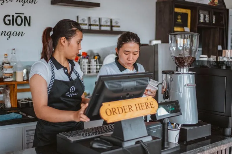 Two women in aprons behind a café counter, one at the till, beneath an “Order here” sign